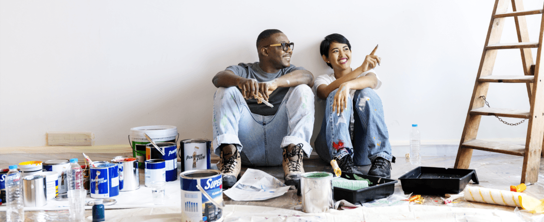 Two people sitting on ground next to ladder and paint buckets