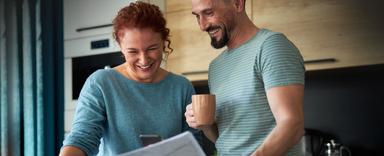 happy couple standing in kitchen holding phone looking at newspaper