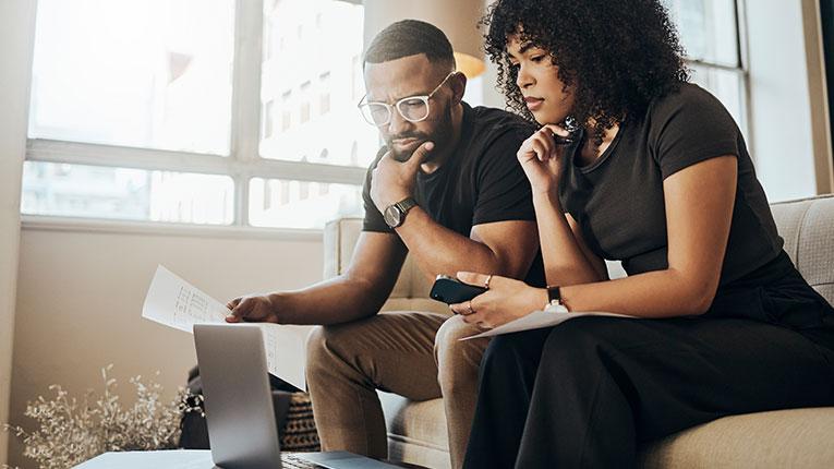 Couple sitting on couch looking worryingly at laptop, holding phone and papers, chins resting on hands