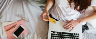 Woman sitting in bed holding credit card with laptop on lap next to phone, notepad and eye glasses