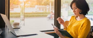 Woman in yellow shirt in coffee shop writing in notebook in front of laptop