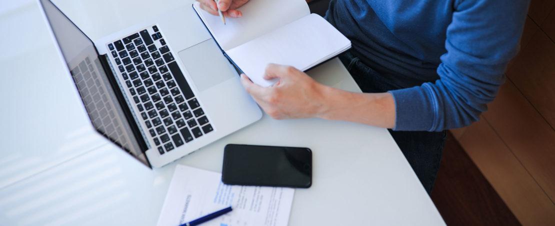 Person in blue shirt sitting at desk with notebook, laptop and phone