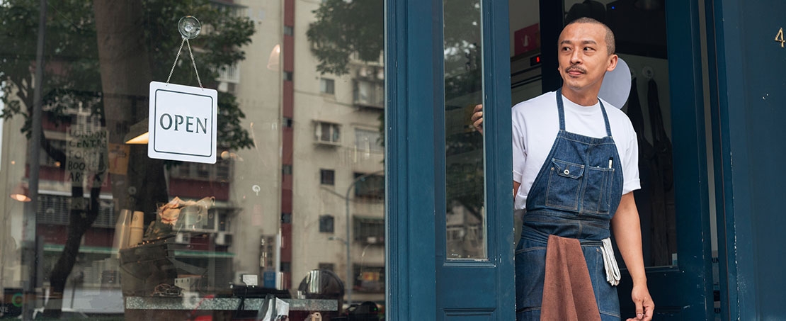 Asian man wearing blue apron looking out shop doorway with sign showing open for business in window